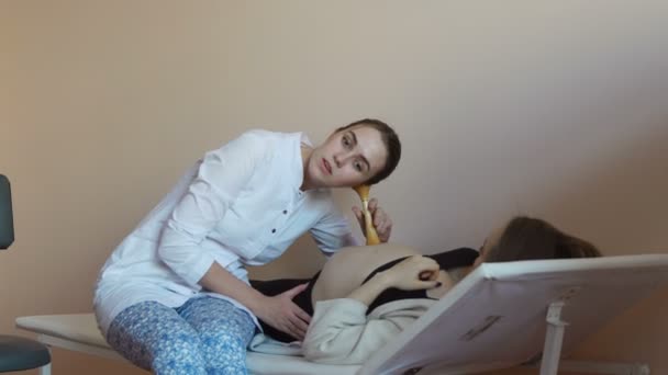 Una donna incinta giace su un divano della clinica e un medico donna ascolta il polso dei bambini attraverso l'addome con l'aiuto di un doppler uditivo e riporta buone notizie. . — Video Stock