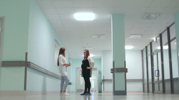 Diálogo entre una mujer embarazada y una doctora en el pasillo del hospital. Preparación para el parto. Consulta sobre el parto. Sonriendo a la gente. Control deslizante. Movimiento horizontal de la cámara — Vídeo de stock