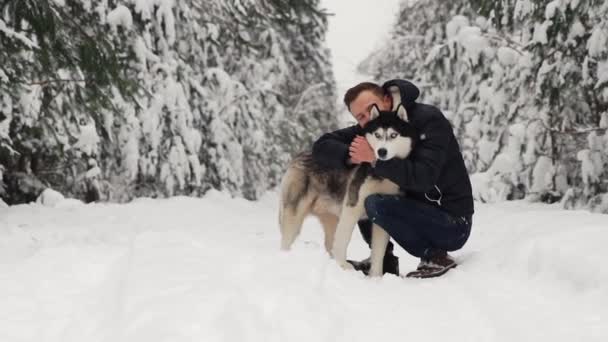 Мужчина обнимает свою сибирскую хаски в лесу. На заднем плане деревья в снегу. Гордость и счастье в глазах. Прогулка с собакой . — стоковое видео