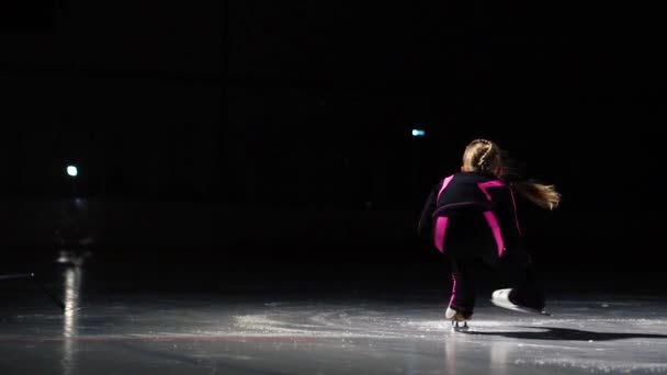 Una piccola pattinatrice in tuta sportiva nera e pattini bianchi esegue una spinning stand nell'arena dello stadio di ghiaccio. Squatting produce una trottola mentre tiene la mano sul bordo del — Video Stock