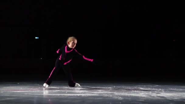 Seorang gadis skater dalam setelan olahraga hitam dan sepatu putih melakukan stand berputar di arena stadion es. Kaki diarahkan ke belakang tangan ke samping. Memutar giroskop — Stok Video