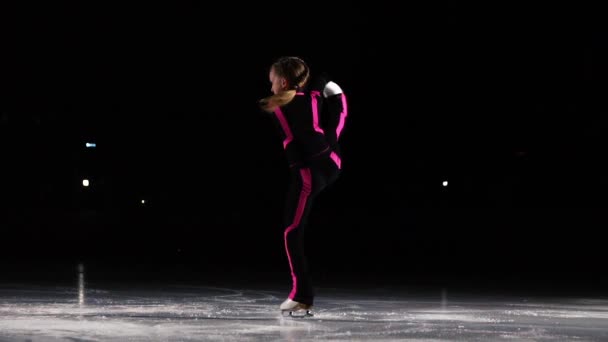 En liten flicka åkare i en svart sport kostym och vita skridskor utför en spinning står på arenan för isstadion. På genomsnittet för konståkning. Stående på ett ben att hålla benet ovan den — Stockvideo