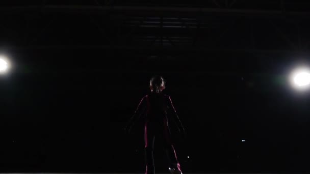 Вид ззаду підсвічування освітлює фігуру професійної дівчини-фігуриста, яка рухається до вогнів. Камера рухається за дівчиною на повзунку. Дівчина в чорному костюмі — стокове відео