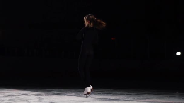 Een vrouwelijke professionele kunstschaatsster in een zwart pak voert de spinnen staande op een been in de positie van een zwaluw op ijs in schaatsen in een slow-motion shot. Arena van de competitie — Stockvideo