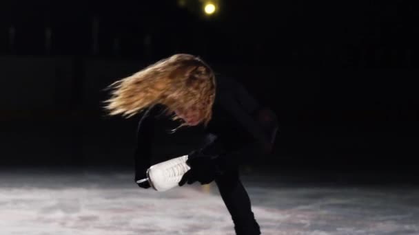 Um close-up de uma garota patinadora no gelo executa uma rotação em torno de seu eixo enquanto está em pé no gelo em uma perna segurando sua mão para a borda do cume. A câmera se move de cima para baixo. O cabelo voa — Vídeo de Stock