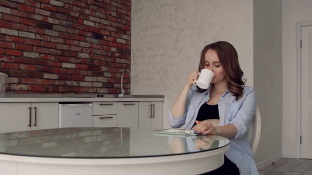 Glad gravid kvinna sitter vid ett glas köksbord dricka kaffe och använder en tablet-dator. gör köp via Internet — Stockvideo