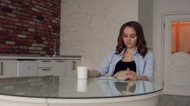 Kahve içme ve bir tablet bilgisayar kullanarak bir cam mutfak masasına oturan mutlu hamile kadın. Yenidoğan için alışveriş online yapar