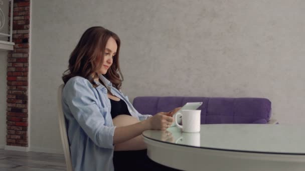 Счастливая беременная женщина сидит за стеклянным кухонным столом, пьет кофе и пользуется планшетным компьютером. Смотрите онлайн лекцию от врача — стоковое видео