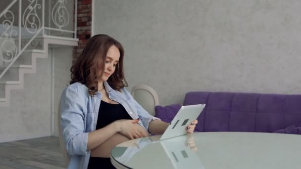 Mulher grávida feliz sentado em uma mesa de cozinha de vidro beber café e usando um computador tablet. Envolvido em negócios na Internet. Trabalho remoto no decreto — Vídeo de Stock