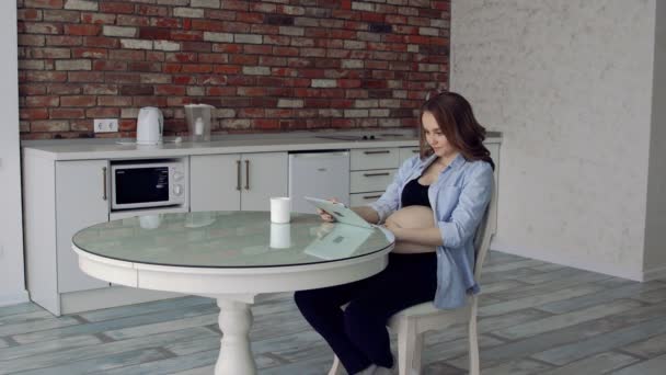 Happy pregnant woman sitting at a glass kitchen table drinking coffee and using the tablet computer. — Stock Video