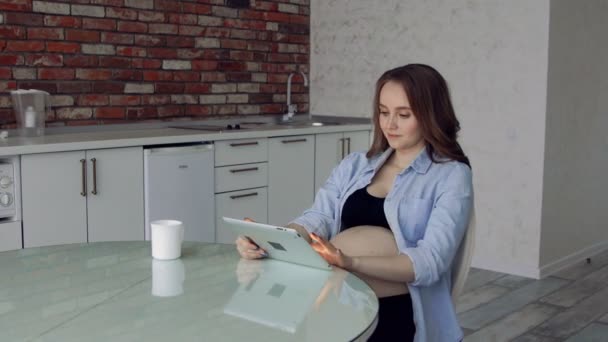 Счастливая беременная женщина сидит за стеклянным кухонным столом, пьет кофе и пользуется планшетным компьютером. делает покупки в Интернете — стоковое видео