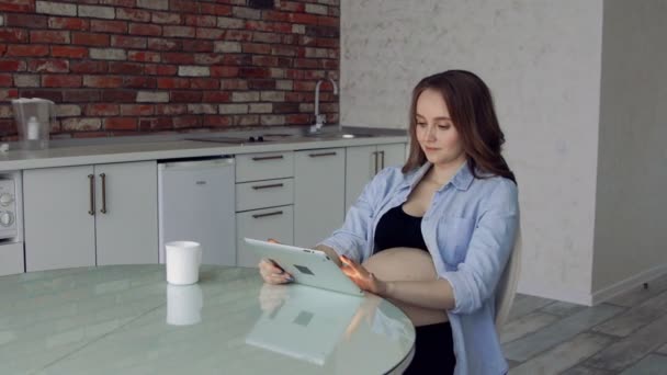 Glad gravid kvinna sitter vid ett glas köksbord dricka kaffe och använder en tablet-dator. gör köp via Internet — Stockvideo