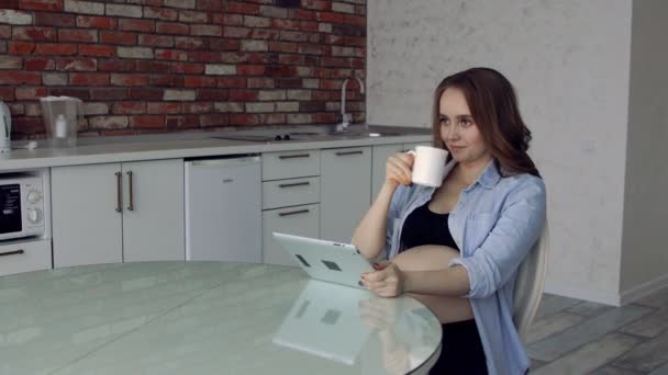 Feliz mujer embarazada sentada en una mesa de cristal de la cocina bebiendo café y usando una tableta. hace compras en línea para recién nacidos — Vídeo de stock