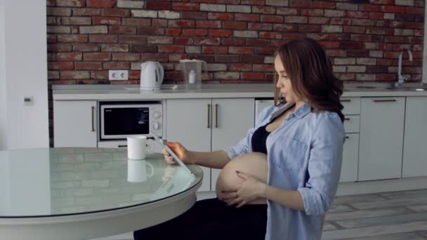 Ευτυχισμένος έγκυος γυναίκα που κάθεται σε ένα τραπέζι κουζίνας γυαλί, πίνοντας καφέ και να χρησιμοποιούν έναν υπολογιστή tablet. Παρακολουθήστε σε απευθείας σύνδεση διάλεξη από τον γιατρό — Αρχείο Βίντεο
