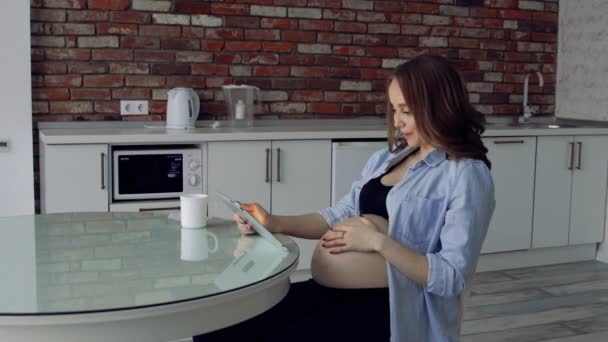 Kahve içme ve bir tablet bilgisayar kullanarak bir cam mutfak masasına oturan mutlu hamile kadın. Aile fotoğrafları izlerken. — Stok video