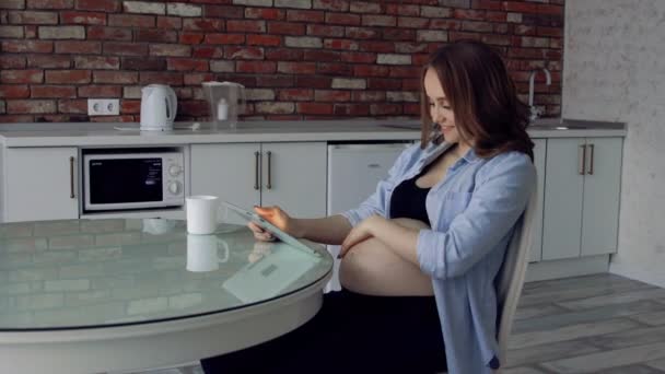 快乐的孕妇坐在玻璃厨房桌子边喝咖啡, 用平板电脑。在互联网上从事业务。遥控工作在旨令 — 图库视频影像