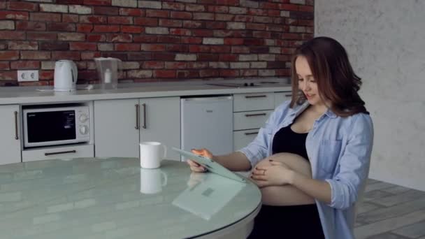 Glad gravid kvinna sitter vid ett glas köksbord dricka kaffe och använder en tablet-dator. Bedriver verksamhet på Internet. Distansarbete är föräldraledig. online video konferens — Stockvideo