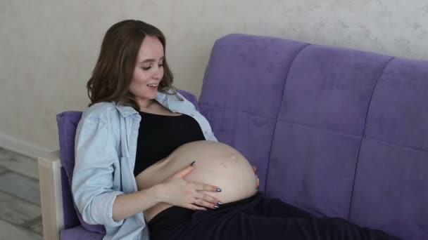 Una mujer embarazada feliz está sentada en el sofá en casa y con la mano acariciando el vientre mientras se comunica con el bebé en el estómago . — Vídeos de Stock