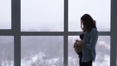 Yüksek bir irtifada bulunan büyük bir panoramik pencere, mutlu bir hamile kadın durumda. Ellerini karnına dokunur ve gülümseyerek karnındaki bebeği ile iletişim kurar. Bakar