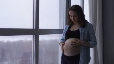 Yüksek bir irtifada bulunan büyük bir panoramik pencere, mutlu bir hamile kadın durumda. O ellerini karnına dokunur ve gülümseyerek bebeğin karnında iletişim kurar.