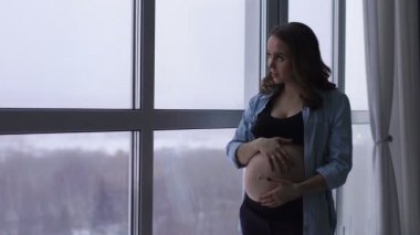 Yüksek bir irtifada bulunan büyük bir panoramik pencere, mutlu bir hamile kadın durumda. Ellerini karnına dokunur ve gülümseyerek karnında bebek ile iletişim kurar. bir portresi