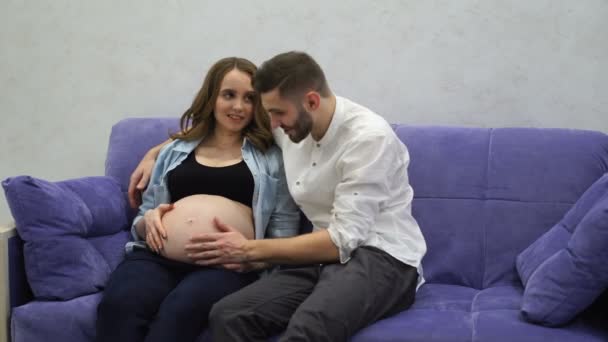 Одружена пара сидить вдома на дивані, чекаючи народження дитини. Чоловік прорвав її руку на живіт і цілує живіт своєї вагітної дружини . — стокове відео
