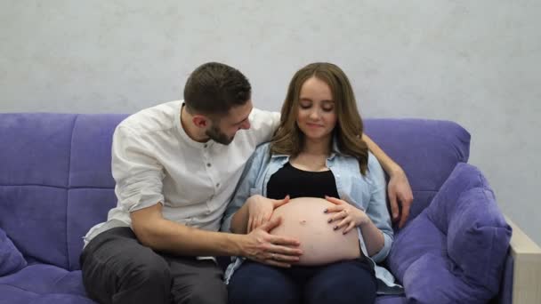 Kobieta w ciąży w domu siedzi na kanapie z mężem oglądania telewizji. Człowiek komunikuje się z żoną i przyszłego dziecka. Całować brzucha i słuchać ruchy dziecka w brzuchu. — Wideo stockowe