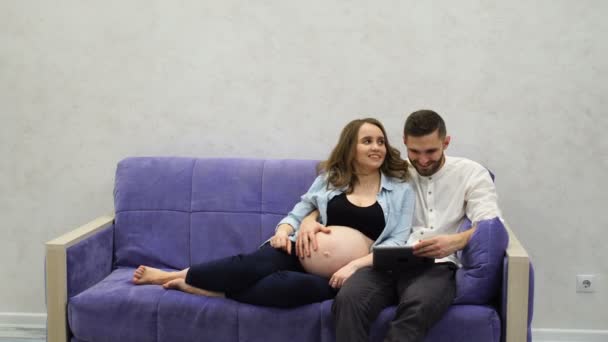 Familia feliz pareja sentada en el sofá llamando a la comunicación de vídeo a sus padres en otra ciudad. Cuenta noticias sobre el embarazo. Una mujer embarazada y un hombre se comunican a través de una tableta a través de — Vídeo de stock