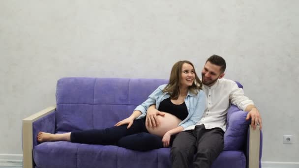 Małżeństwo w domu siedzi na kanapie, czeka na narodziny dziecka. Człowiek gładził jej rękę na jej brzuch i Całowanie brzucha żony w ciąży. — Wideo stockowe