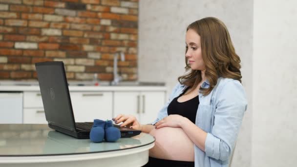 Una mujer embarazada escribe en un foro de madres con su computadora portátil sentado en una mesa de vidrio. Salón interior blanco. Comunicación por Internet de las mujeres embarazadas . — Vídeos de Stock