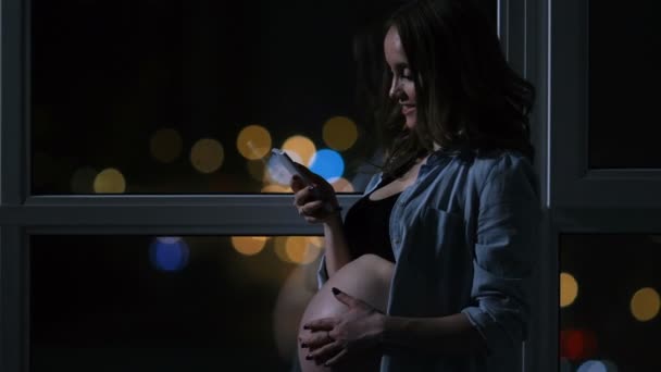 Femme enceinte la nuit parlant sur un téléphone portable touchant son estomac debout à la grande fenêtre panoramique donnant sur la ville. Les voitures passent par la fenêtre. La fille regarde dans l'écran du téléphone — Video