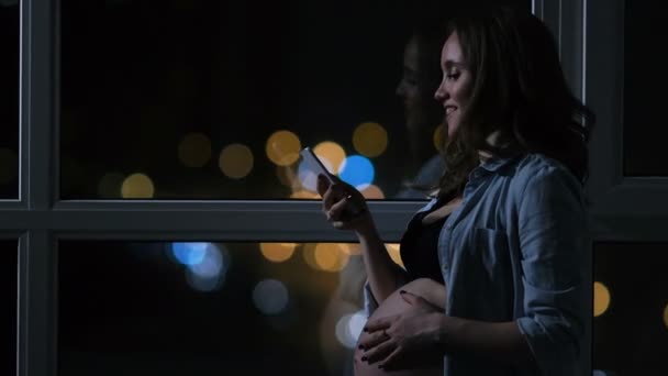 Mujer embarazada por la noche hablando en un teléfono móvil tocando su estómago de pie en la gran ventana panorámica con vistas a la ciudad. Los coches pasan por la ventana. La chica mira la pantalla del teléfono — Vídeo de stock