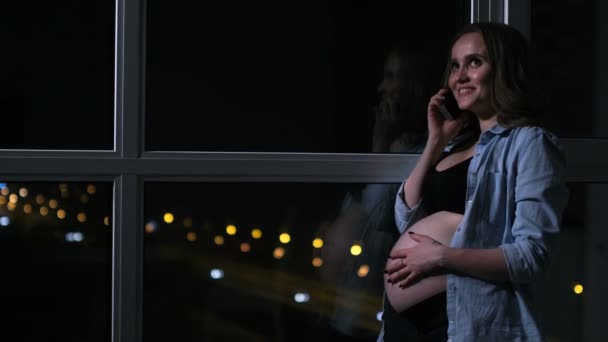 Беременная женщина в ночи разговаривает по мобильному телефону руки касаются желудка стоя у окна — стоковое видео