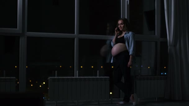 Una mujer embarazada por la noche hablando en un teléfono móvil tocándose el estómago con las manos de pie en la gran ventana panorámica con vistas a la ciudad . — Vídeos de Stock