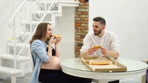 Hombre y mujer embarazada comiendo pizza en casa en su cocina. Una comida rica en grasas, hambrienta — Vídeo de stock