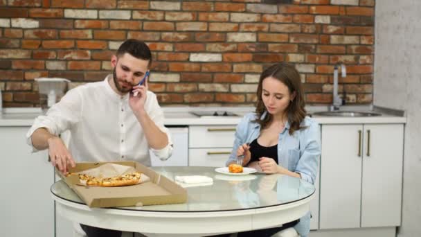 Homem e mulher grávida comendo pizza em casa em sua cozinha. Homem falando ao telefone durante o jantar — Vídeo de Stock