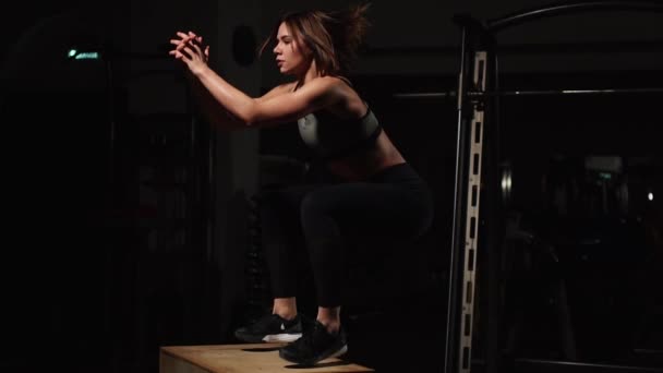 Belle athlète de fitness féminine effectue des sauts de boîte dans une salle de gym sombre portant un haut de sport noir et collants courts avec le visage caché — Video