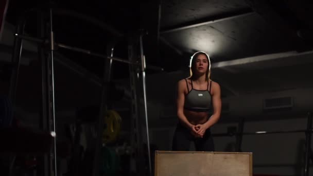 Hermosa atleta de fitness femenina realiza saltos de caja en un gimnasio oscuro con top deportivo negro y medias cortas con la cara oculta — Vídeos de Stock