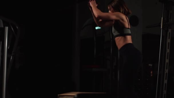 美丽的女性健身运动员表演方块跳跃在黑暗的健身房穿着黑色运动顶部和短紧身衣与脸隐藏 — 图库视频影像