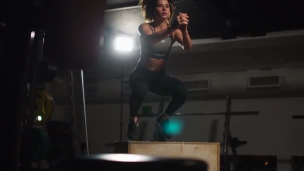 Beautiful female fitness athlete performs box jumps in a dark gym wearing black sports top and short tights with face hidden — Stock Video