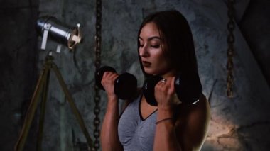 Kısa şort ve taş gri bir duvar arka plan üzerinde kapalı dumbbells ile şişirilmiş basın seksi sportif kız ellerini pazı eğitim için dumbbells ile yükseltir. Yakın çekim
