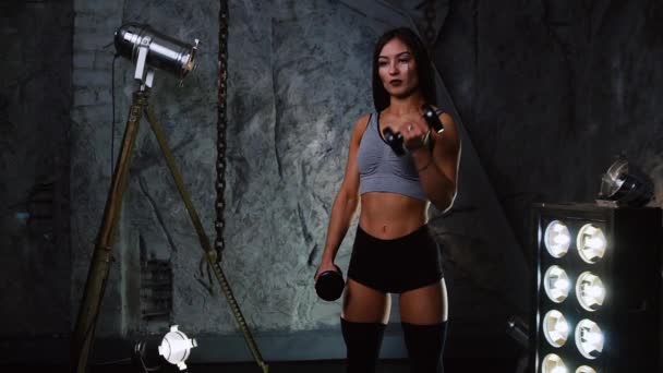 Dívka sexy sportovní s činkami v místnosti v pozadí šedé kamenné zdi účastní školení zbraní — Stock video