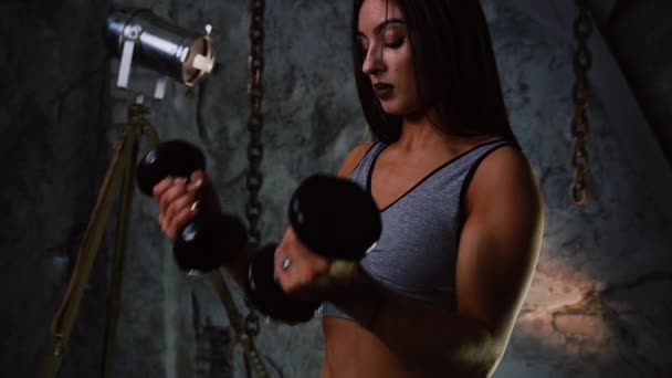 Sexy chica deportiva en pantalones cortos y prensa inflada con mancuernas en el interior en el fondo de una pared gris piedra levanta las manos con mancuernas para el entrenamiento del bíceps. De cerca. Levantamiento simultáneo de — Vídeos de Stock