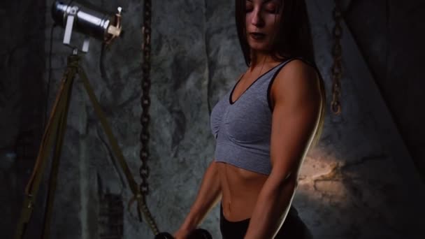 Sexy sporty girl with a beautiful inflated booty and inflated press with dumbbells on the background of a stone gray wall raises her hands with dumbbells for bicep training. Close up. Beautiful girl — Stock Video