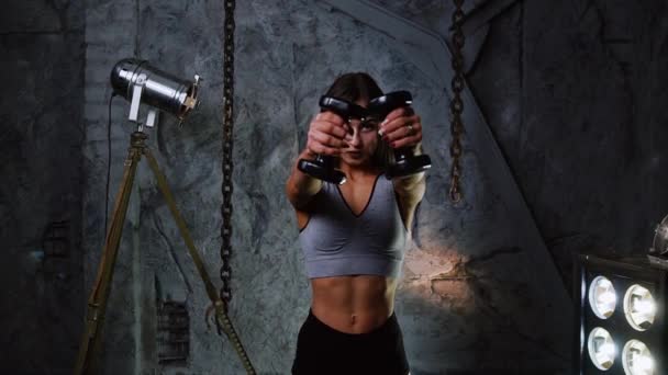 Спортивная девушка в сексуальной одежде смотрит прямо в камеру и делает упражнения на плечах с гантелями. Красивые надутые мышцы живота — стоковое видео