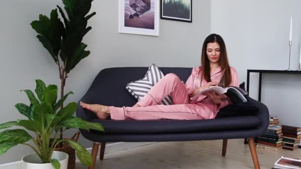 Uma linda garota de pijama rosa sexy lê uma revista brilhante deitada no sofá e bebendo champanhe de um copo . — Vídeo de Stock