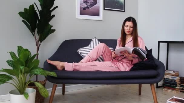 Una hermosa chica en pijama rosa sexy lee una revista brillante tumbada en el sofá y bebiendo champán de una copa. En el salón de su casa — Vídeos de Stock