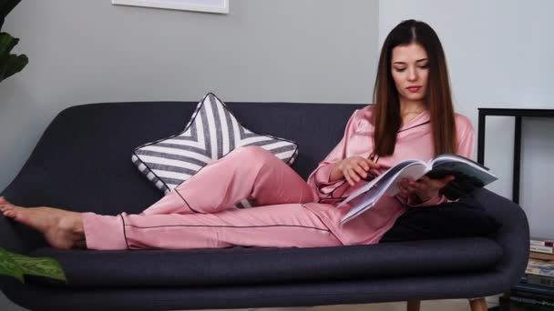 Uma linda garota de pijama rosa sexy lê uma revista brilhante deitada no sofá e bebendo champanhe de um copo. Na sala de estar da casa dele. Ler revistas de moda — Vídeo de Stock