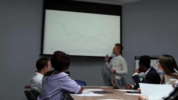 Un joven empleado de la empresa en una camisa blanca informa sobre el progreso del trabajo realizado durante el último año y los planes de desarrollo para el ajetreado trimestre en el diagrama del proyector en el — Vídeos de Stock