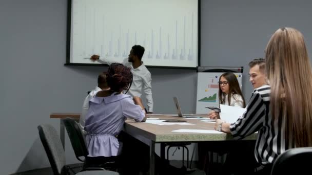 Afrika kökenli Amerikalı antrenör ofiste iş adamları için kurumsal tanıtım sağlar, koyu tenli işadamı yeni iş planı üzerinde beyaz tahta, istemcilere toplantısında hoparlör dinleme konuşma sunar — Stok video